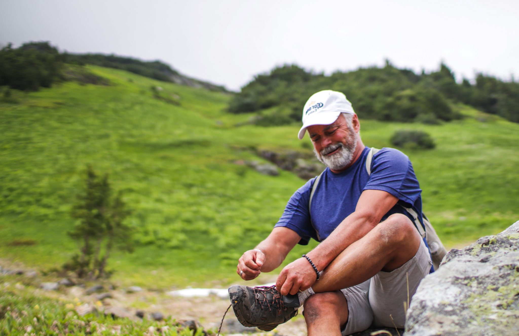 A middle aged man pauses to tie his shoe on a hike in a hilly, green landscape